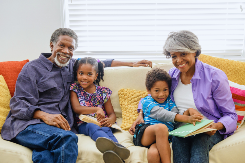 Grandparents sitting with their two grandchildren on a couch at home, looking at books together