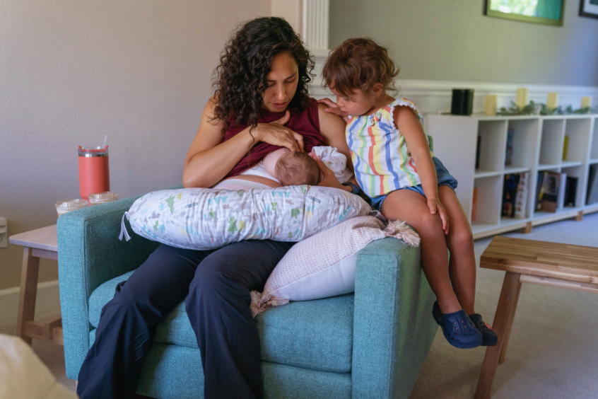 A mother breastfeeding her newborn baby while her toddler daughter is looking at the baby curiously